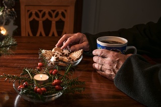 Hands of an elderly single man sitting alone at a table with Christmas cookies, coffee and festive decoration next to an empty chair, lonely holidays during the croronavirus pandemic or after a loss, selected focus