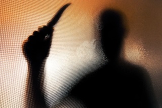 Colour backlit image of the silhouette of a man wielding a sharp knife in an aggressive way. The silhouette is distorted, and the arms elongated, giving an alien-like quality. The image is sinister and foreboding, with an element of horror. The image conveys a domestic violence, knife crime theme. Horizontal image with copy space.