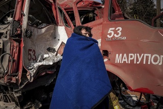 Žena sa po ostreľovaní v Mariupole zakrýva dekou v blízkosti poškodeného hasičského auta.
