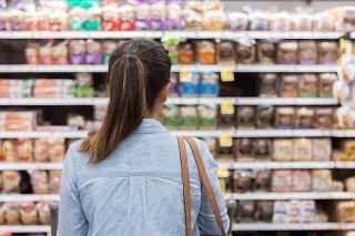 In this rear view, an unrecognizable woman stands with a shopping cart in front of a shelf full of food in the bread aisle of a grocery store.