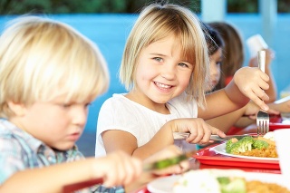 Elementary Pupils Enjoying Healthy Lunch In Cafeteria Sitting Down Smiling At Camera