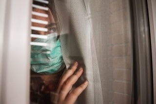 A young woman in quarantine wearing a mask and looking through the window