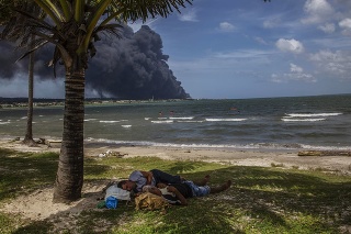 Dvaja muži spia pri palme a ďalší ľudia sa kúpu na pláži neďaleko požiaru skladiska paliva v provincii Matanzas na Kube.