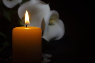 A close up of an orange candle and flame and lily flowers on a dark background.
