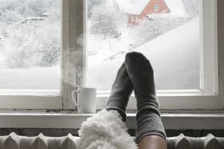 Cozy winter still life: woman legs in warm woolen socks under shaggy blanket and mug of hot beverage on old windowsill against snow landscape from outside.