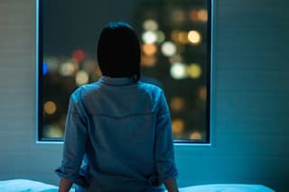 A rear view of a woman sitting alone on a bed in room and looking through the window at night.