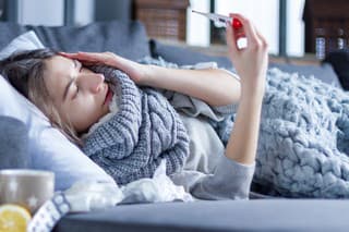 Sick exhausted girl in scarf is lying in bed wrapped in blanket. Young woman with fever and headache is measuring temperature with thermometer, treated at home. Winter cold and flu concept.