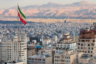Waving Iran flag above skyline of Tehran at sunset.