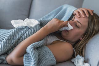 Sick Woman having Flu with Caught Cold. Sneezing into Tissue. Headache. Virus. Sick woman lying in bed with high fever. She is blowing nose. Photo of Young female Lying On Bed covered with a blanket  Infected With Allergy holding her head.