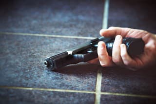 A hand lying on a tiled floor clasps a semi-automatic pistol. The owner may be dead or injured, and the incident may have been accidental, deliberate, or have happened in the course of a crime.