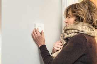 A woman dressed in a warm wool jumper and scarf, adjusting a home thermostat dial.