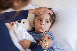 Sick boy with thermometer laying in bed and mother hand taking temperature. Mother checking temperature of her sick son who has thermometer in his mouth. Sick child with fever and illness while resting in bed.
