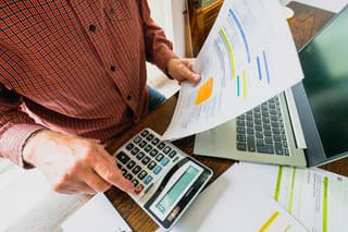 Wide angle image depicting a senior man's hand holding an energy bill while the other hand checks the numbers on a calculator. The table is strewn with documents and also a laptop. Room for copy space.