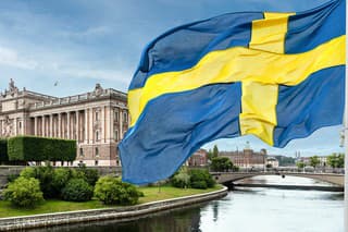 The building of the Swedish Parliament (Riksdag) and the Riksbank Bridge over the Lilla Vartan Strait with the national flag of Sweden in the foreground.