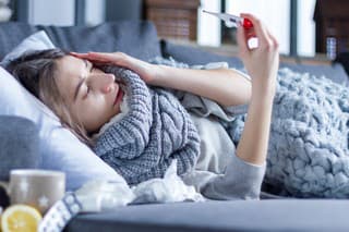 Sick exhausted girl in scarf is lying in bed wrapped in blanket. Young woman with fever and headache is measuring temperature with thermometer, treated at home. Winter cold and flu concept.