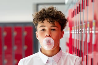A waist-up shot of a male teenager blowing a bubble with bubble gum standing next to red lockers in school between lessons. He is looking into the camera.