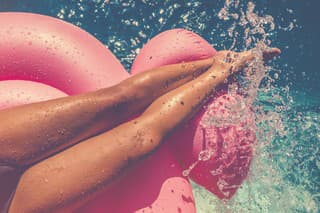 Woman floating on a pink inflatable in swimming pool. Tight crop showing only her legs and feet. She is tanned in turquoise water with water splashing onto her.