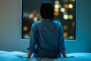A rear view of a woman sitting alone on a bed in room and looking through the window at night.