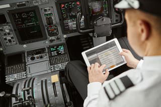 Male pilot use digital tablet in passenger airplane jet. Interior of cockpit in modern plane with dashboard and air navigation. Top view of man wear uniform. Civil aviation. Air travel concept