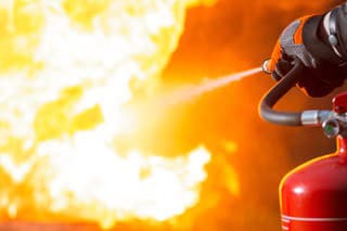 Close-up showing a person pointing the nozzle of a fire extinguisher at the base of a large fire.