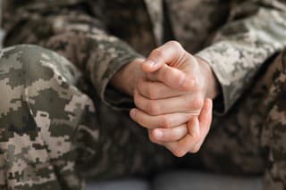 Cropped of soldier sitting on couch alone, suffering from loneliness and depression at home after returning from military service, hands of unrecognizable man in camouflage uniform, closeup