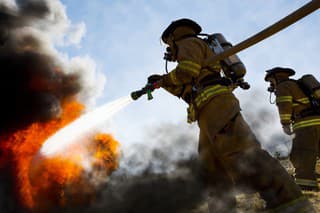 Firefighters in a fire protection suit wearing firefighter helmet with breathing device and holding fire hose is extinguishing a burning house fire that is putting off excessive heat and smoke.  Fire could have been caused by accident or arson.  Second firefighter in background.  