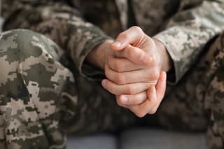 Cropped of soldier sitting on couch alone, suffering from loneliness and depression at home after returning from military service, hands of unrecognizable man in camouflage uniform, closeup