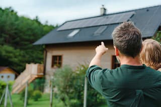Rear view of dad holding her little girl in arms and showing at their house with solar panels.Alternative energy, saving resources and sustainable lifestyle concept.