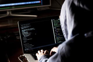 hacker in hoodie typing code in program using laptop in dark studio