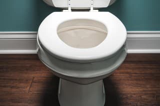 A elongated white toilet with the seat lid in the up position. The toilet is highlighted with a spot light coming from above. This sparse room has blue-green walls and a dark hardwood floor.