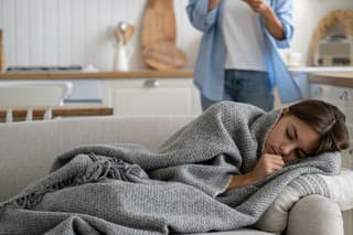 Sick child girl having fever and flu symptoms lying on sofa under blanket while her worried stressed mother talking to doctor by phone, calling to pediatrician, selective focus. Telehealth concept