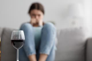 Sad caucasian millennial female sits on sofa suffering from stress and troubles, focus on bottle and glass of wine in home interior. Alcoholism, drinking alone, depressed, addicted to alcohol at home
