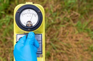 A blue gloved hand holds a radiation meter. The meter shows a high level of radiation. Grass in the background, most of which is brown and dead.
