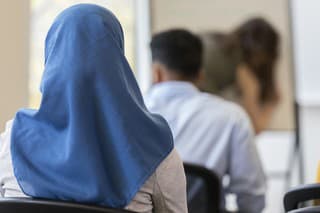 In this closeup rear view, an unrecognizable woman wearing a hijab sits in a chair in a classroom.  Other classmates sit in front of her.
