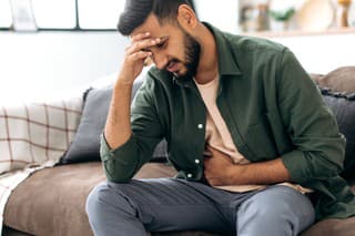 Unhappy indian or arabian man, sits on comfortable sofa in cozy living room, holds his hands on his stomach, grimaces from pain in his stomach, suffers from poisoning, spasm, stomach problems