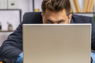 Confident young businessman hiding behind laptop computer, looking at camera, spying his colleagues working, peeping. Professional freelancer man looking from behind computer with cunning eyes glance