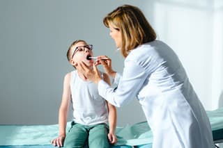 Female doctor checking a sore throat of her young male patient wearing eye glasses. He is sitting on the bed while the doctor is using tongue depressor to have a better look at his throat.