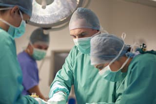 a female operating nurse stands over a patient at the operating table and looks down to what she is doing in the operation. She is joined by a young female nurse and mature male surgeon .In the background the anaesthetist is looking over from his monitors .  They are all wearing surgical gowns and protective masks and headwear. they are wearing green gowns . The shot is horizontal waist up , with defocussed background for copy space .