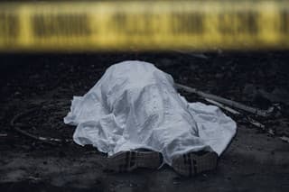 The dead body is seen lying on the ground behind a cordon tape.