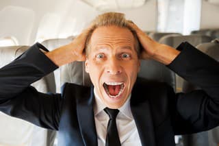 Shocked mature businessman touching his head with hands and looking at camera while sitting at his seat in airplane