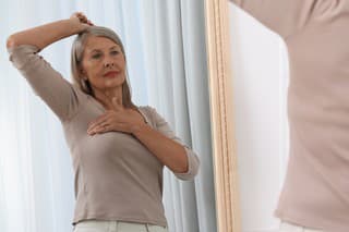 Beautiful senior woman doing breast self-examination near mirror indoors