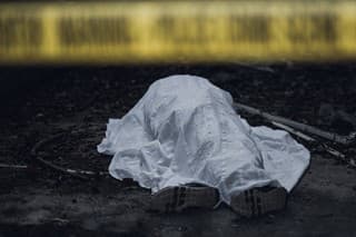 The dead body is seen lying on the ground behind a cordon tape.