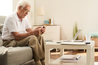 Portrait of modern senior man learning to use smartphone and laptop at home in cozy living room