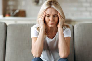 Middle aged blonde woman sits on couch at living room holding her head with her hands, feels unhappy because of headache, personal troubles, illness or bad news, she need psychological or medical support