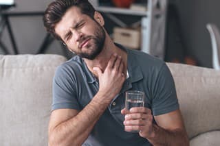 Frustrated handsome young man touching his neck and holding a glass of water while sitting on the couch at home