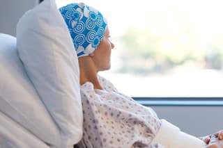 Young woman in bed suffering from cancer. Thoughtful woman battling with tumor looking out of window. Young patient with blue headscarf recovery in hospital on bed.