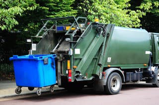 Green garbage truck with an elevated blue wheelie bin at the rear