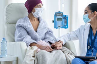 A senior woman battling cancer sits in a high back chair as she talks with her Oncologist during a Chemotherapy treatment.  The patent is dressed casually in a sweater and has a blanket and headscarf on to keep her warm.