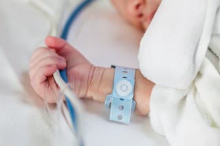 Close-up of a caucasian newborn baby holding life-support hoses and cables tight in his had with wrist nametag while sleeping in a hospital bed.