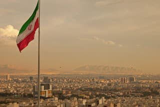 Iran flag waving  in the wind above skyline of Tehran lit by orange glow of sunset.